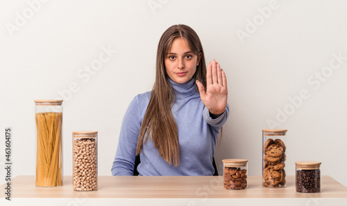 Young caucasian woman sitting at a table with food pot isolated on white background standing with outstretched hand showing stop sign, preventing you.
