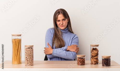 Young caucasian woman sitting at a table with food pot isolated on white background frowning face in displeasure, keeps arms folded.
