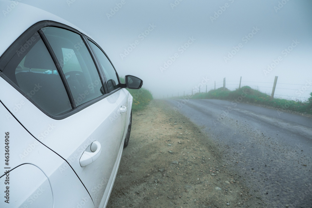 Side of a white car parked off road. Small asphalt road in a country side in a fog. Dark and moody pastel color. Nobody. Dangerous driving conditions concept. Low visibility