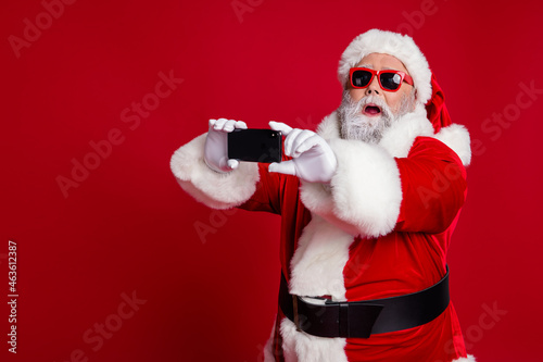 Portrait of attractive bearded amazed funny Santa using device taking making selfie vlog isolated over bright red color background
