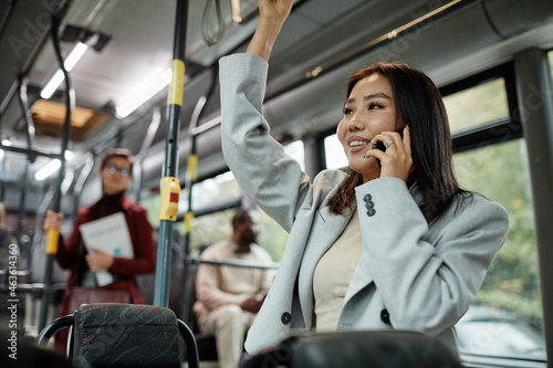 Portrait of smiling Asian woman speaking by smartphone in bus and holding onto handle while traveling by public transport in city, copy space
