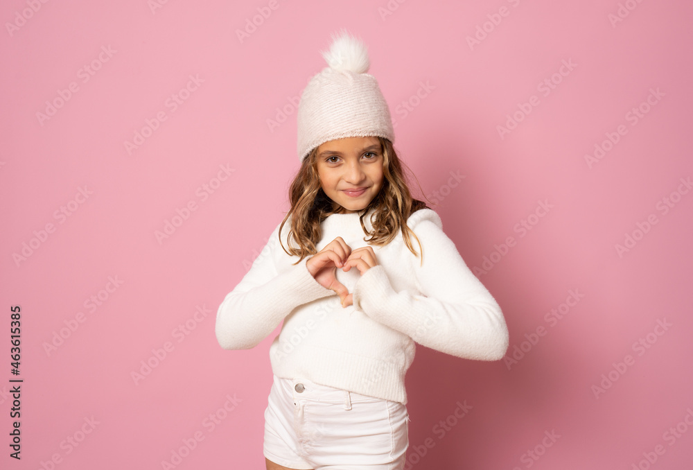 little girl in white winter knitted hat and sweater making heart figure on pink background isolated.