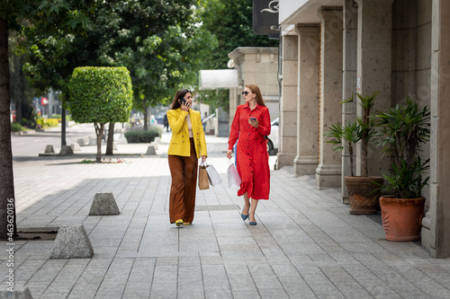 Two women walking down the street after shopping carrying their bags and talking on the phone