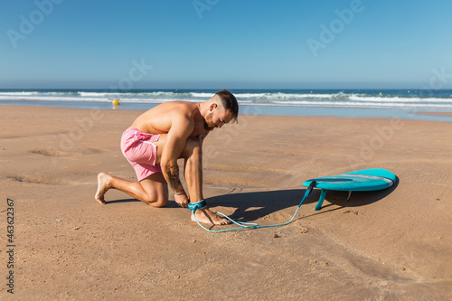 Sportive surfer attaching leash to leg on shore