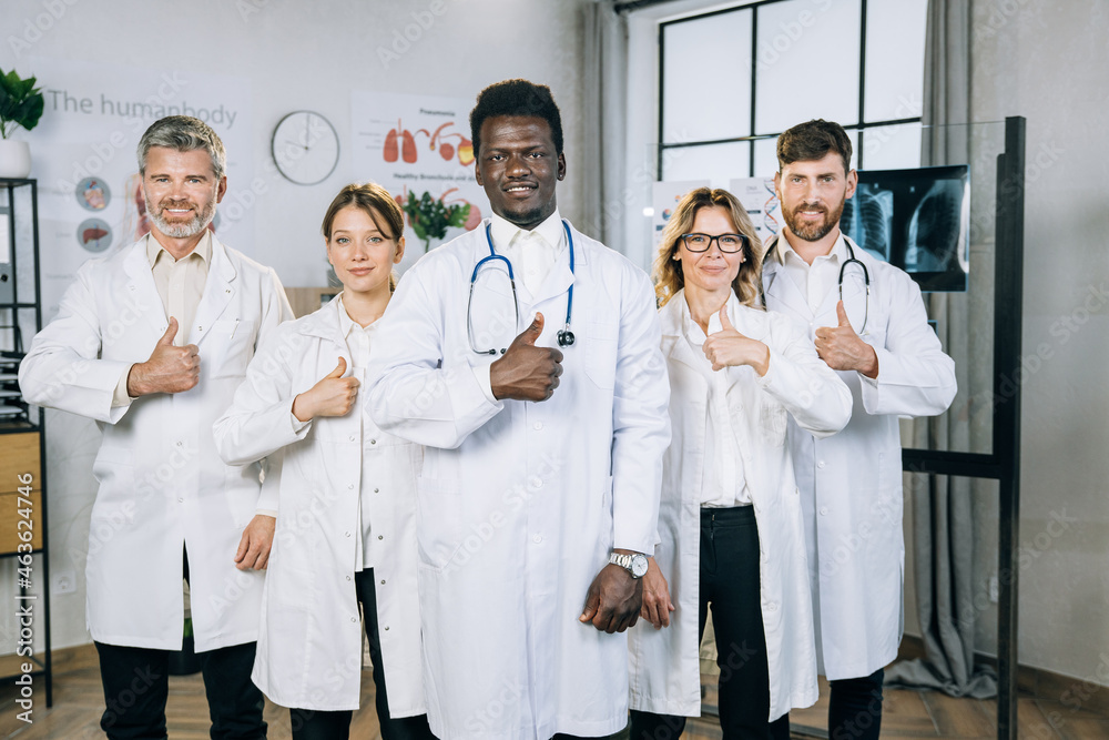 Portrait of happy five multiracial doctors in medical uniform standing at office room, showing their thumbs up and smiling on camera. Concept of people, medicine and health care. Portrait of happy five multiracial doctors in medical uniform standing at office room, showing their thumbs up and smiling on camera. Concept of people, medicine and health care.