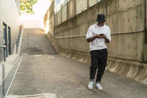 Chico negro atlético posando con el smartphone en una calle gris con ropa urbana