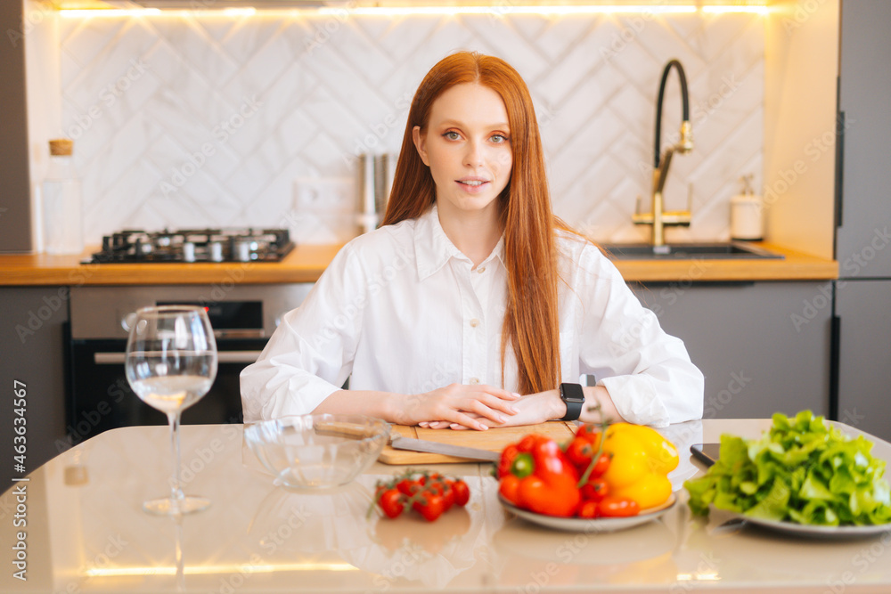 Medium shot portrait of attractive redhead young woman sitting at kitchen table with cutting board, knife and fresh vegetables, looking at camera. Beautiful female cooking vegetarian meals at home.