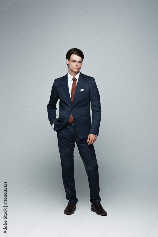 full length of young androgynous businessman in suit posing with hand in pocket on grey full length of young androgynous businessman in suit posing with hand in pocket on grey