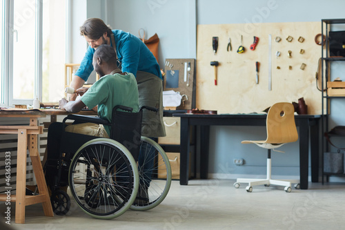 Full length side view portrait of young man in wheelchair learning leathermaking craft in leatherworkers workshop, copy space photo