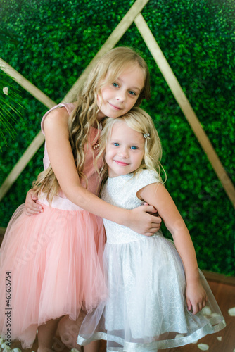 Little cute girls sisters with blond hair in a white and pink dresses and white flowers, lilies and orchids on a background with a green tropical plants