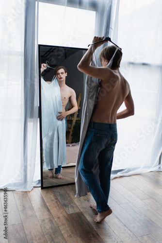 full length of young transgender man with makeup holding hanger with slip dress near mirror