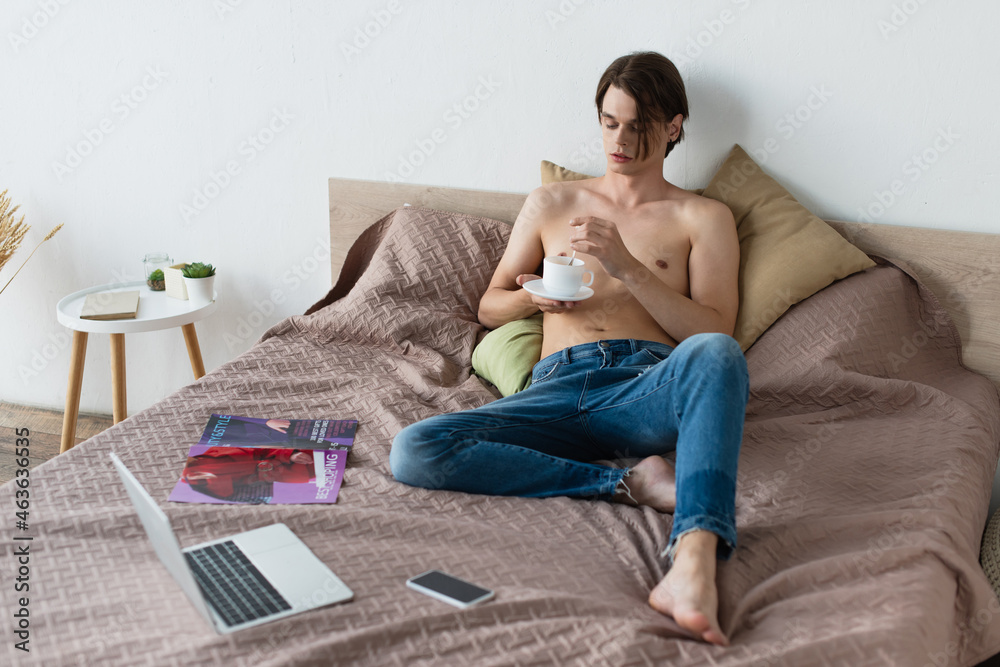 barefoot transgender young man in jeans holding cup of coffee near gadgets on bed