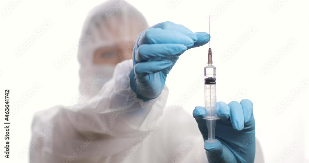 Doctor in a protective suit with a syringe looking to a syringe with vaccine. Blurred background. Concept of health care, and infection and virus protection. Doctor in a protective suit with a syringe looking to a syringe with vaccine. Blurred background. Concept of health care, and infection and virus protection.