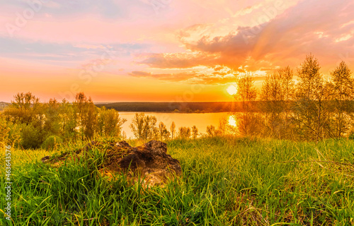 Scenic view at beautiful sunset on a shiny lake with old rough stone on the foreground, green grass, birch trees, golden sun rays, calm water ,nice cloudy sky on a background, spring landscape