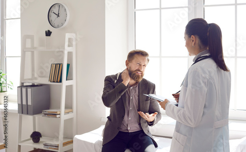 Male patient talking to doctor about pain in the nape of his neck. Female general practitioner listening to man who can't stretch stiff tight injured neck due to severe fibromyalgia or muscle strain photo