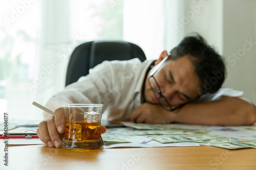 Stressed adult Asian businessman at workplace and drinking alcohol with many document paper for business on desk. Asian man overstressed at job while sitting at office desk in the office photo