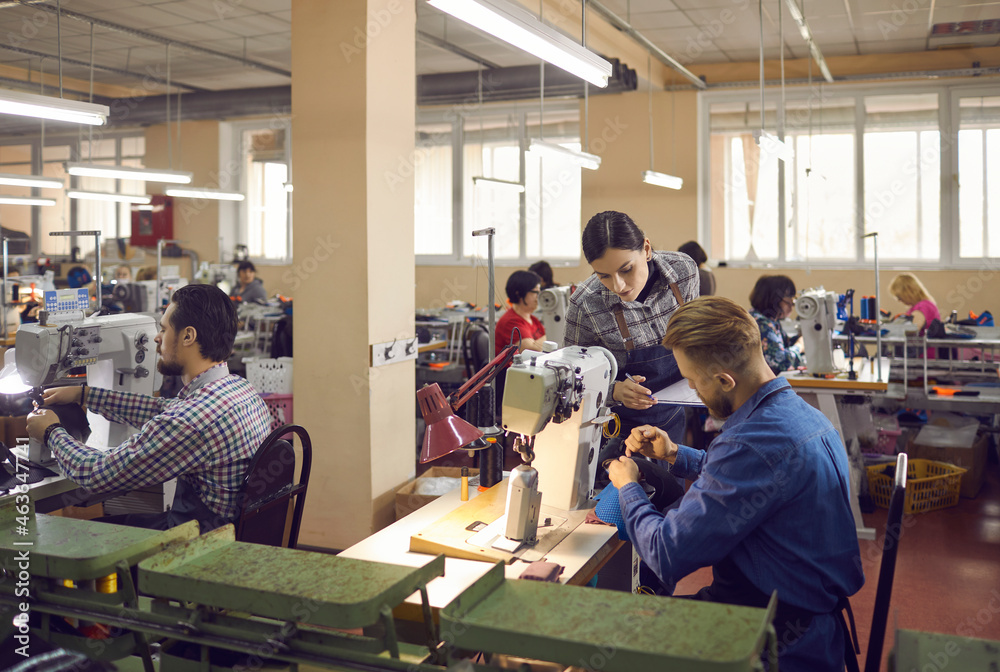 People working in a big workshop room at a shoe factory. Male and female workers sitting at tables with industrial sewing machines and making new footwear details. Manufacturing industry concept - obrazy, fototapety, plakaty 