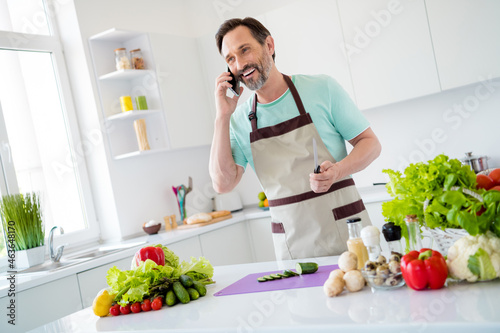 Photo of charming cute age man dressed blue t-shirt apron cooking talking modern device smiling indoors home room