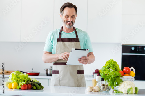 Photo of modern mature man hold tablet choose recipe typing wear apron blue t-shirt home kitchen indoors