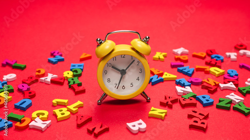 Random colorful alphabet and yellow alarm clock on a red background, colorful letters. Top view.