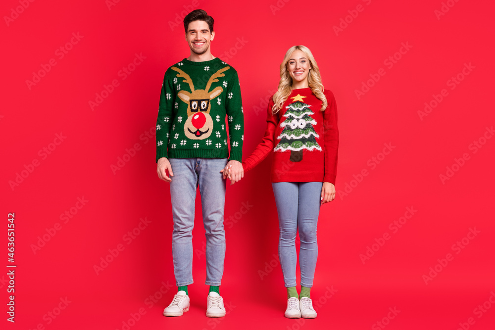Photo of cheerful positive couple hold hands toothy smile wear ugly sweater jeans shoes isolated red color background Photo of cheerful positive couple hold hands toothy smile wear ugly sweater jeans shoes isolated red color background