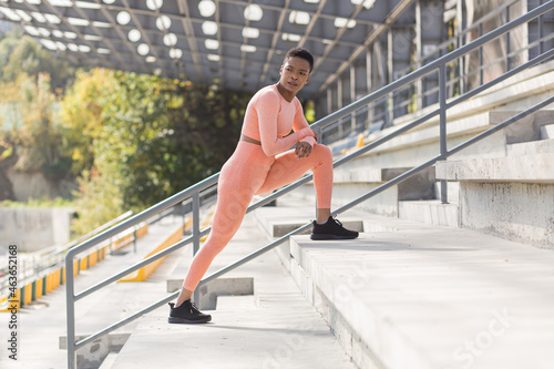 Female athlete doing fitness in a pink sports suit near the sports stadium, African American performs fitness exercises and stretching in the morning © Liubomir