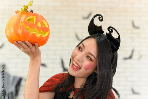 Portrait of young beautiful asian woman wearing witch costume holding pumpkin Jack O Lantern on white brick background decorated with black paper bat shape for halloween party