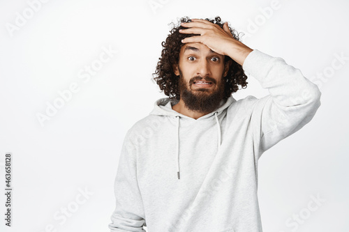 Yikes. Shocked young man slap forehead, looking troubled, forgot smth, panicking, standing in hoodie over white background photo