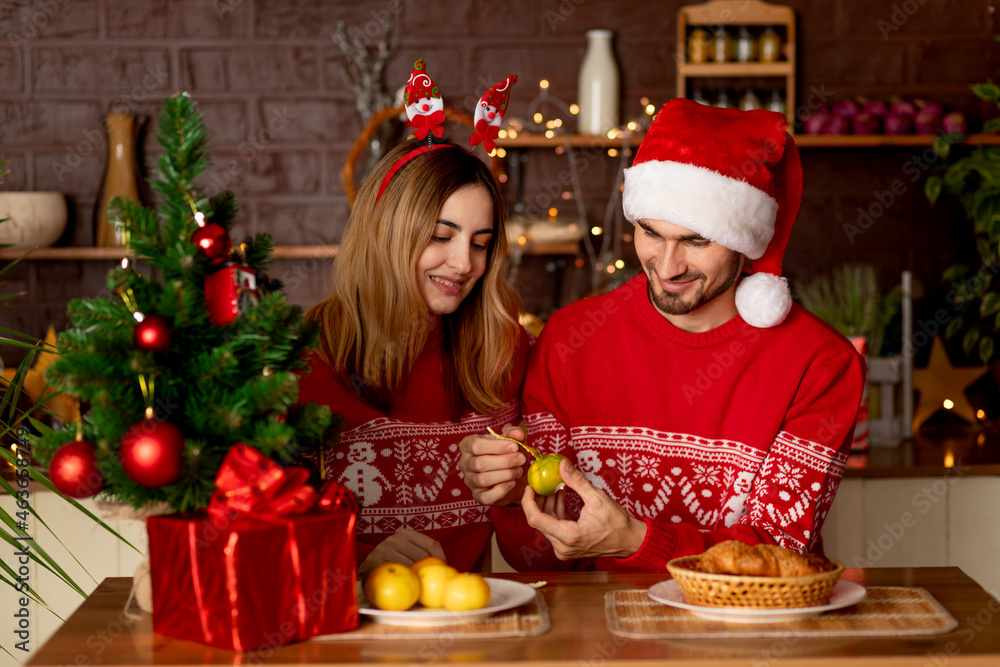 happy lovers a man and a girl in red sweaters in the kitchen with a Christmas tree feed each other tangerines at home and celebrate New Year or Christmas rejoicing and smiling happy lovers a man and a girl in red sweaters in the kitchen with a Christmas tree feed each other tangerines at home and celebrate New Year or Christmas rejoicing and smiling