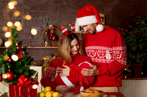 a happy young family mom dad and baby in red sweaters in the kitchen with a Christmas tree at home celebrate New Year or Christmas rejoicing in the gift of the birth of a child