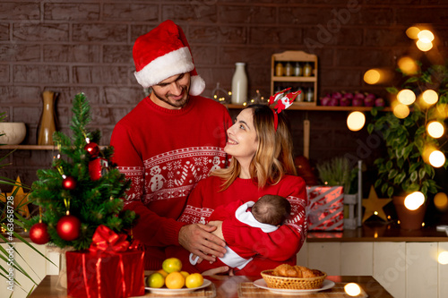 a happy young family mom dad and baby in red sweaters in the kitchen with a Christmas tree at home celebrate New Year or Christmas rejoicing in the gift of the birth of a child