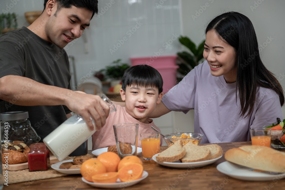 Happy Asian family enjoying breakfast together on dinning table. Young Asian father pouring a glass of milk to his son while having breakfast in the kitchen. Happy Asian family lifestyle concept.