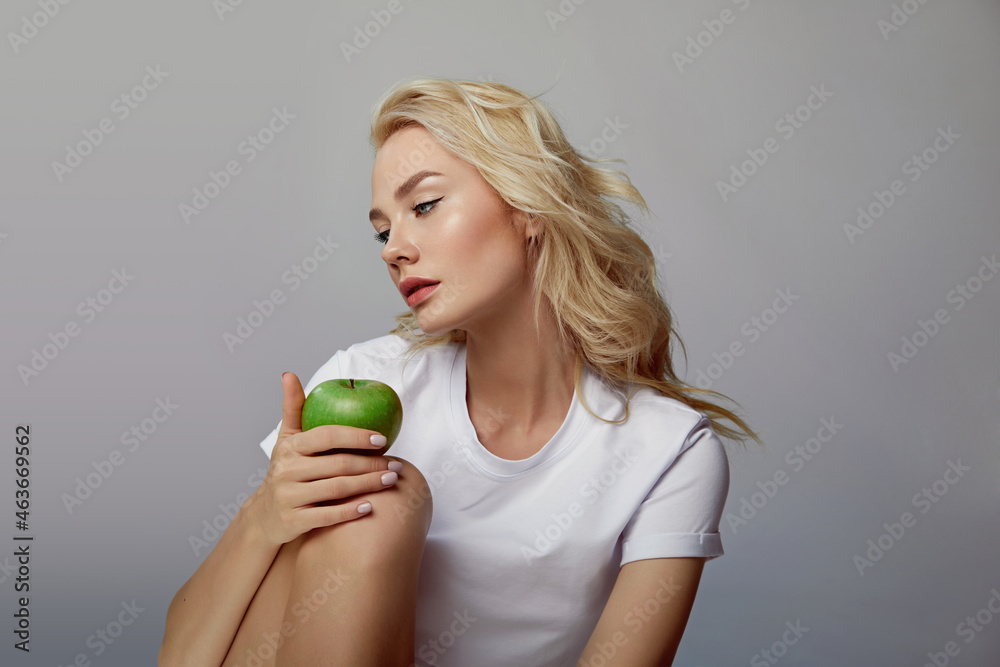portrait of a beautiful young woman with blond hair and blue eyes in a white t-shirt. Woman holding a green apple near her face. Natural food concept. Natural cosmetics. portrait of a beautiful young woman with blond hair and blue eyes in a white t-shirt. Woman holding a green apple near her face. Natural food concept. Natural cosmetics.