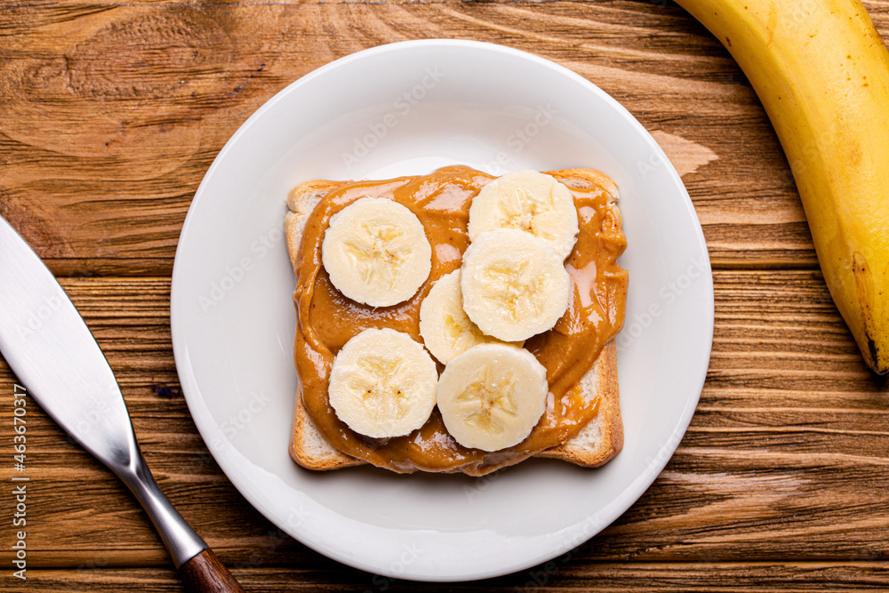 Toast with peanut butter and banana on white ceramic plate with knife on rustic wooden background,  jar with spoon, top view flat lay