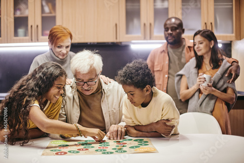 Grandchildren playing board game together with their grandfather at the table at home with parents standing in the background