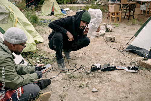 Two young male refugees using smartphones while sitting on ground in their camp