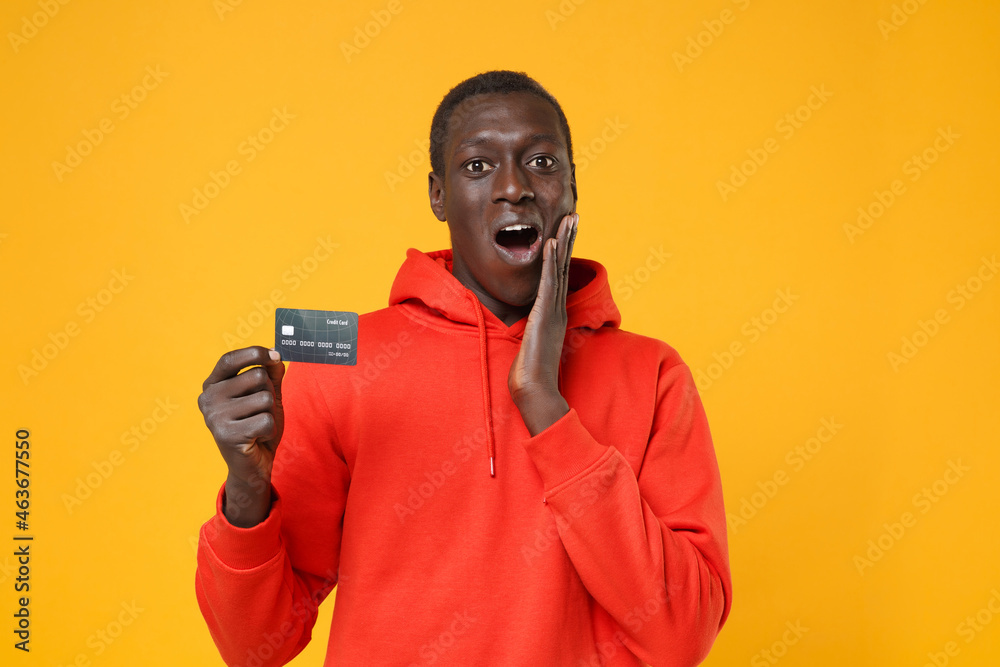 Shocked young african american man guy in red streetwear hoodie posing isolated on yellow background in studio. People lifestyle concept. Mock up copy space. Hold credit bank card, put hand on cheek. Shocked young african american man guy in red streetwear hoodie posing isolated on yellow background in studio. People lifestyle concept. Mock up copy space. Hold credit bank card, put hand on cheek.