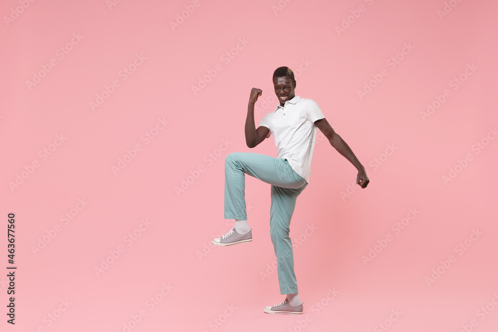 Joyful young african american man guy in white polo shirt, turquoise trousers posing isolated on pastel pink background in studio. People lifestyle concept. Mock up copy space. Doing winner gesture.