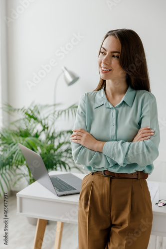 Young smiling successful employee business woman in casual blue shirt work look aside stand hold hands crossed at workplace desk with laptop pc computer at office indoors. Achievement career concept. © ViDi Studio