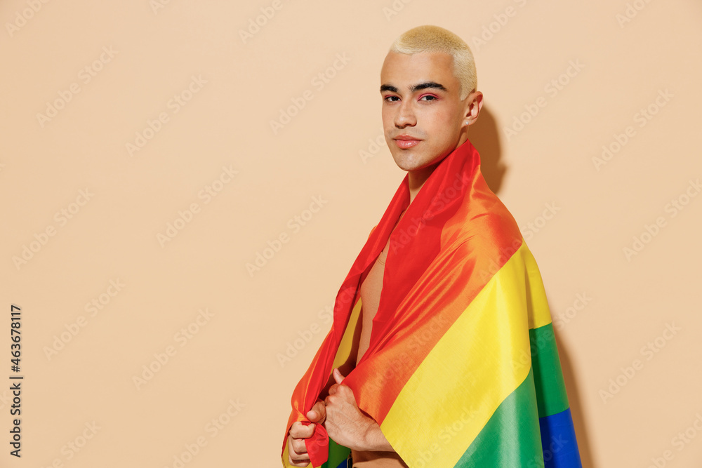 Side view young confident happy blond latin gay man with make up in beige tank shirt wrapped in rainbow flag look camera isolated on plain light ocher background studio People lgbt lifestyle concept. Side view young confident happy blond latin gay man with make up in beige tank shirt wrapped in rainbow flag look camera isolated on plain light ocher background studio People lgbt lifestyle concept.