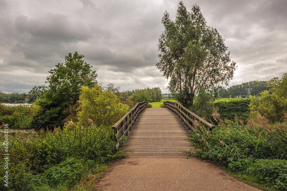 Wooden bridge over the Dutch river Dommel. The photo was taken on a cloudy day at the end of the summer season near the village of Raamsdonksveer, province of North Brabant. Fall is coming already. Wooden bridge over the Dutch river Dommel. The photo was taken on a cloudy day at the end of the summer season near the village of Raamsdonksveer, province of North Brabant. Fall is coming already.