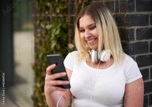 Happy young cheerful millennial student girl standing in front of a black brick wall and chatting on her smartphone with headphones on her neck - People lifestyle and technology concept with teenager