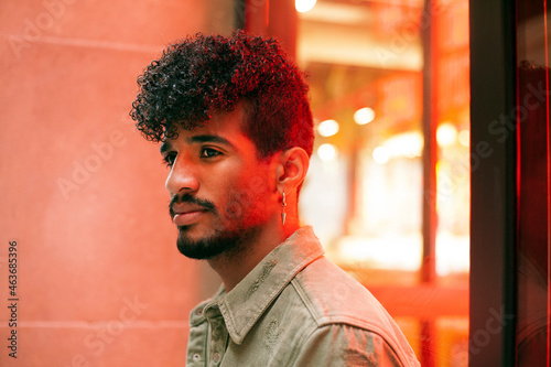 Portrait of young biracial curly haired man with orange neon light reflection on face. Looking to the side. Millennial with beard and earring. Urban style. Horizontal image. photo