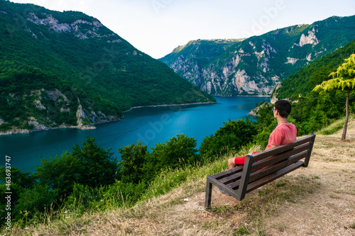 Guy tourist sits on a bench against the backdrop of river canyon and Lake Piva located between high mountain ranges near Pluzine. Montenegro.