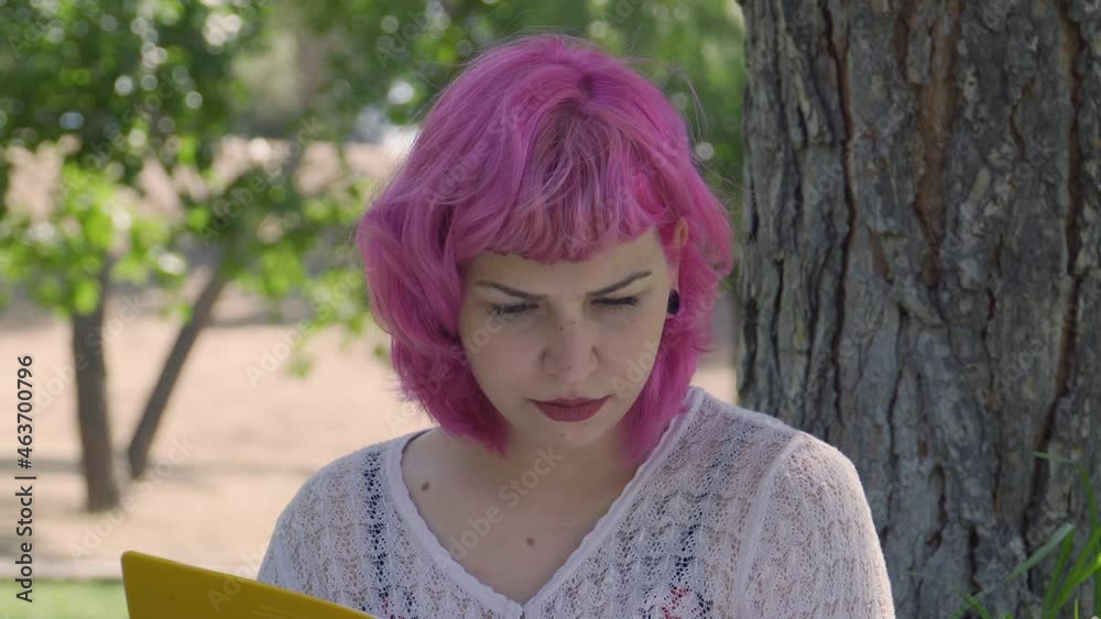 Mujer de pelo rosa leyendo un libro y pasando las páginas  , mujer de pelo corto y labios rojos leyendo junto a un árbol , chica joven estudiando en el parque, universitaria estudiando al aire libre