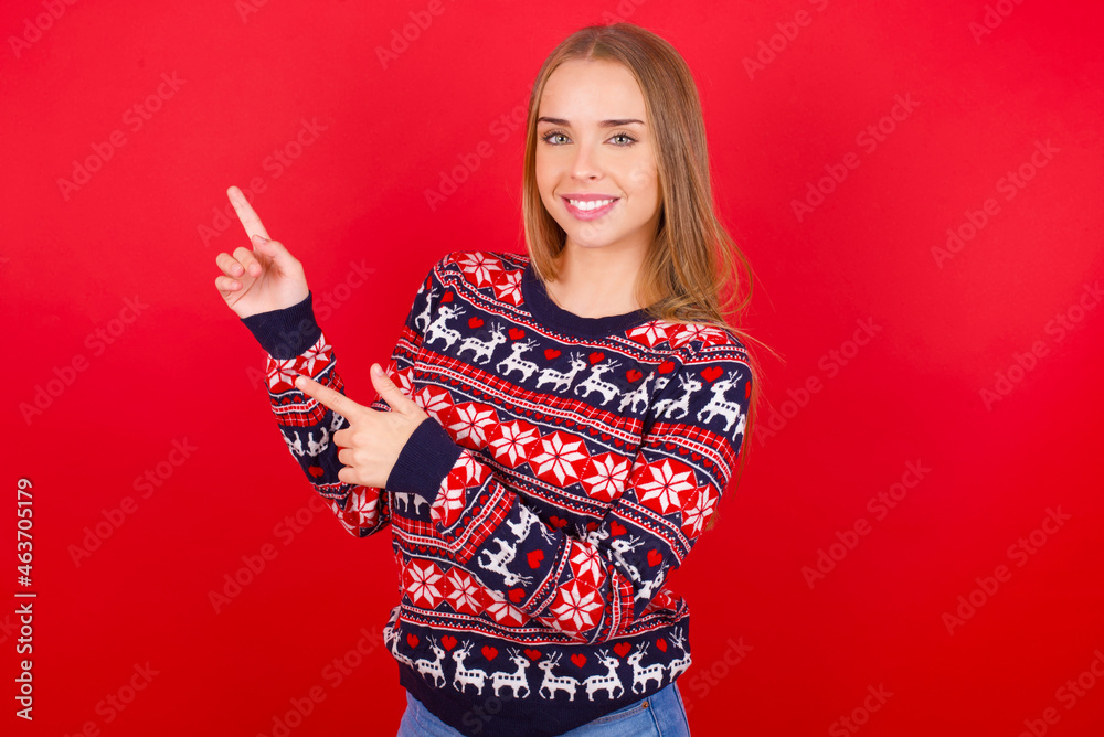 Young caucasian girl wearing christmas sweaters on red background indicating finger empty space showing best low prices, looking at the camera Young caucasian girl wearing christmas sweaters on red background indicating finger empty space showing best low prices, looking at the camera