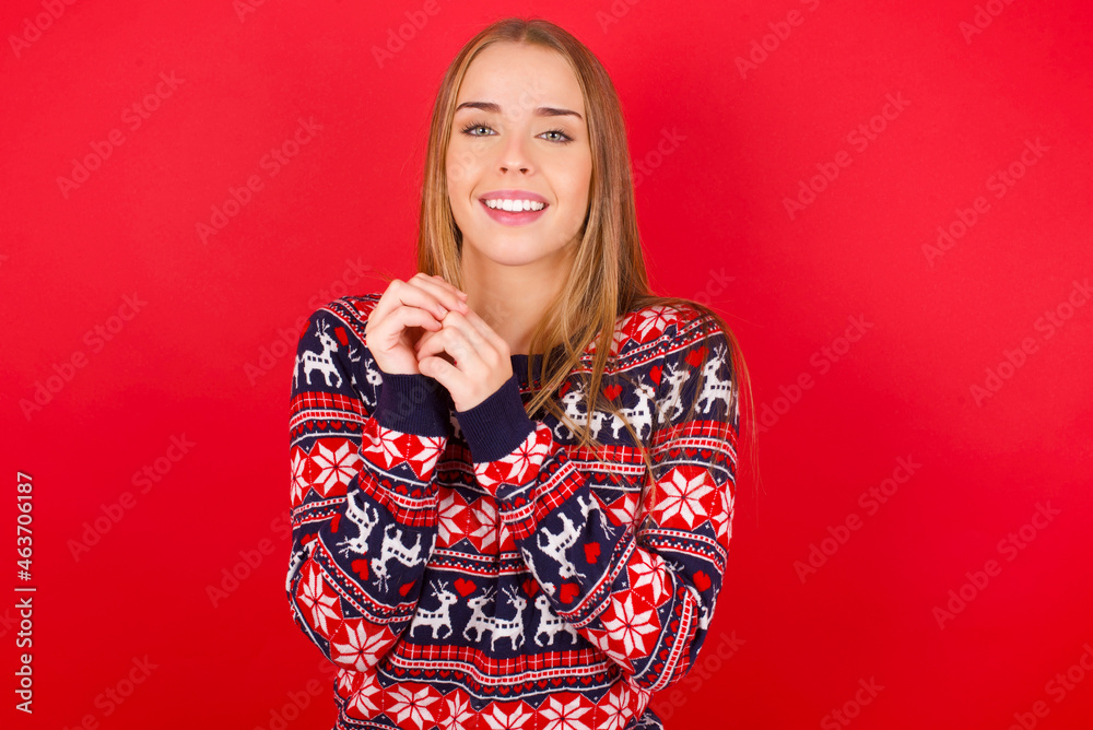 Positive Young caucasian girl wearing christmas sweaters on red background smiles happily, glad to receive pleasant news from interlocutor, keeps hands together. People emotions concept. Positive Young caucasian girl wearing christmas sweaters on red background smiles happily, glad to receive pleasant news from interlocutor, keeps hands together. People emotions concept.