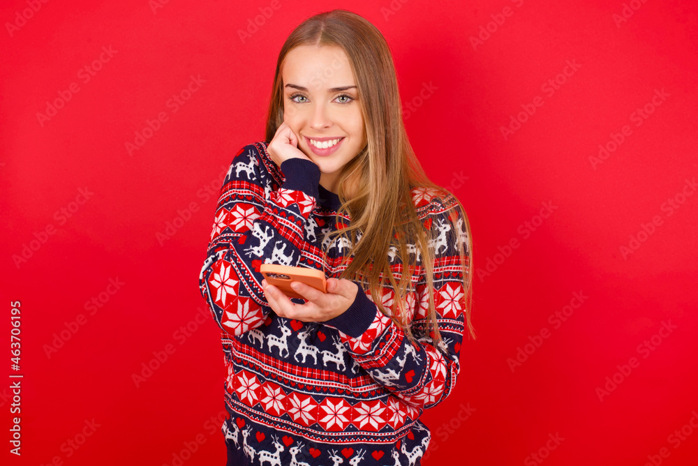 Joyous Young caucasian girl wearing christmas sweaters on red background poses with mobile phone device, types text message on modern smartphone, watches funny video during free time,