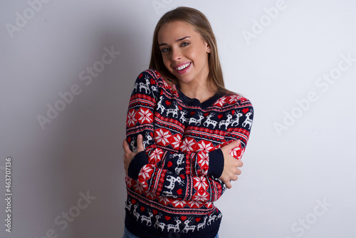 Charming pleased Young caucasian girl wearing christmas sweaters on white background embraces own body, pleasantly feels comfortable poses. Tenderness and self esteem concept