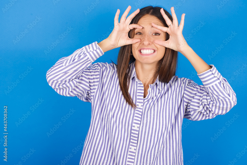 Young european woman wearing striped shirt on blue bakcground keeping eyes opened to find a success opportunity. Young european woman wearing striped shirt on blue bakcground keeping eyes opened to find a success opportunity.